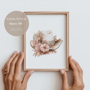 Könnte beinhalten: Ein gerahmtes Kreuzstichmuster-PDF mit einem Cowboyhut, der mit rosa Blumen und Weizen geschmückt ist. Der Hut ist cremefarben mit einem dekorativen Band. Der Text "CROSS STITCH Pattern PDF" befindet sich in einem Kreis oben links.