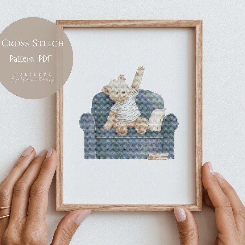 May include: A cross stitch pattern PDF featuring a light brown teddy bear wearing a white striped shirt, sitting on a blue armchair with one arm raised. The bear is next to a small white pillow and a stack of books, all framed in a light wood frame.
