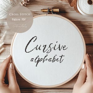 May include: A cross-stitch design featuring the words "Cursive alphabet" stitched in black thread on white fabric, held in a wooden embroidery hoop. The image includes text that reads "Cross Stitch Pattern PDF" and the seller's name.