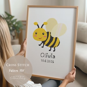 May include: A framed cross-stitch pattern showing a cartoon bee with yellow and black stripes, pink cheeks, and a smiling face. The name "Olivia" and the date "10.6.2025" are below the bee. The frame is light brown.