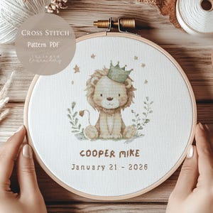 May include: A cross-stitch pattern in a wooden hoop features a lion wearing a crown, surrounded by stars and foliage. The text "COOPER MIKE" and "January 21 - 2026" are stitched below the lion. The words "CROSS STITCH PATTERN PDF" are also visible.