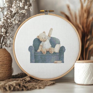 May include: A circular wooden embroidery hoop containing white fabric with a cross-stitch design of a light brown teddy bear wearing a white and grey striped shirt. The bear is sitting on a blue armchair with one arm raised, next to a small pillow.
