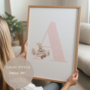 Könnte beinhalten: Ein gerahmtes Kreuzstichmuster mit einem großen rosa Buchstaben "A" mit weißen Punkten. Ein kleiner Hase steuert ein rosa Flugzeug. Der Text "CROSS STITCH Pattern PDF" und "Juliette Embroidery" sind ebenfalls sichtbar.