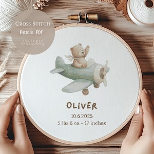May include: Cross-stitch pattern PDF in a wooden hoop. The design features a teddy bear in a light blue airplane. The name "OLIVER", date, weight (5 lbs 6 oz), and height (17 inches) are stitched below the plane.