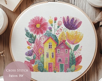 May include: A cross-stitch pattern featuring two colorful houses surrounded by flowers and foliage in a wooden embroidery hoop. The design includes pink, yellow, and purple flowers, with the text "Cross Stitch Pattern PDF" on a circular label.