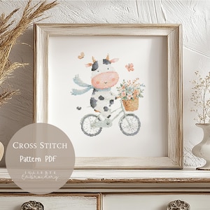 Puede incluir: Un patrón de punto de cruz enmarcado que representa una vaca en bicicleta, con una cesta de flores. La vaca es blanca y negra con una cara rosa y una bufanda azul. El marco es de color marrón claro. Texto: "Cross Stitch Pattern PDF".