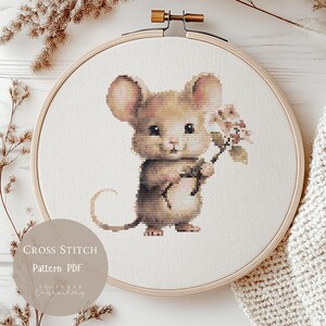 Puede incluir: Patrón de punto de cruz de un ratón adorable con flores, en un bastidor de bordado de madera. El ratón es en tonos marrones y beige, con detalles florales. El texto "Cross Stitch Pattern PDF" es visible.