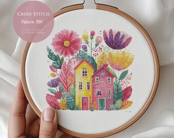 May include: Cross-stitch pattern featuring two colorful houses surrounded by flowers and foliage in a wooden embroidery hoop. The design includes pink, yellow, purple, and green elements. Text on the image reads "Cross Stitch Pattern PDF".