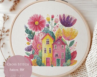 May include: A cross-stitch pattern in a wooden embroidery hoop. The design features two colorful houses surrounded by flowers and foliage in shades of pink, yellow, purple, and green. The text "Cross Stitch Pattern PDF" is visible.