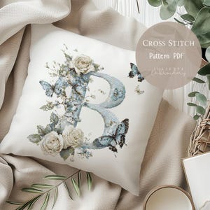 May include: A white pillow with a cross-stitch design featuring the letter "B" in light blue, adorned with butterflies and cream-colored roses. The pillow has the text "Cross Stitch Pattern PDF" and "Juliette Embroidery".