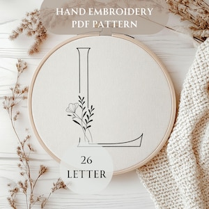 Könnte beinhalten: Ein weißer Stickrahmen mit einer schwarzen Umrandung des Buchstabens L und einem kleinen Blumenmuster. Der Rahmen ist mit "26 BUCHSTABE" beschriftet.