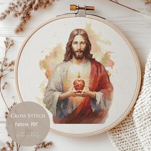 Op de afbeelding: Een kruissteekpatroon van Jezus die een hart vasthoudt, omlijst in een houten borduurring. De afbeelding heeft een zacht kleurenpalet met tinten rood, beige en bruin. De tekst "Cross Stitch Pattern PDF" is zichtbaar.