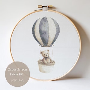 Puede incluir: Patrón de punto de cruz en un bastidor de madera, con un globo aerostático con un oso de peluche en una cesta. El globo está a rayas en gris y crema. El texto "Cross Stitch Pattern PDF" está en la parte inferior.