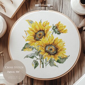 May include: A cross-stitch pattern of sunflowers in a wooden embroidery hoop. The sunflowers are yellow and green, with detailed stitching. The text "CROSS STITCH Pattern PDF" is visible on the bottom left. The background is a wooden surface.