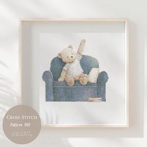 May include: A cross stitch pattern featuring a light brown teddy bear wearing a white striped shirt, sitting on a dark blue armchair with one arm raised. A small white pillow and a stack of books are placed on the seat next to the bear.