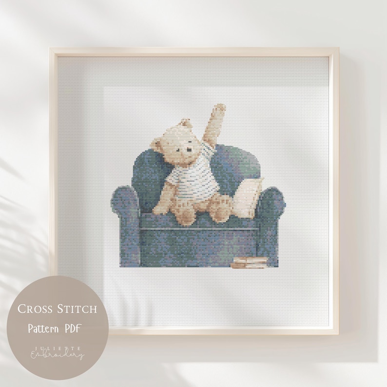May include: A cross stitch pattern featuring a light brown teddy bear wearing a white striped shirt, sitting on a dark blue armchair with one arm raised. A small white pillow and a stack of books are placed on the seat next to the bear.