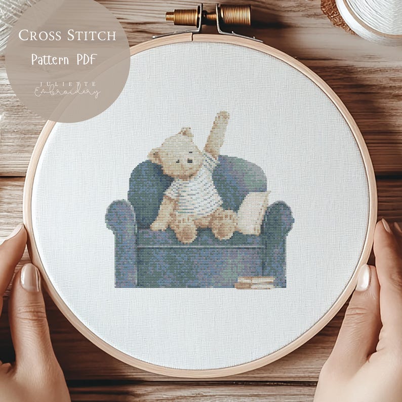 May include: A cross stitch pattern featuring a light brown teddy bear wearing a white striped shirt, sitting on a dark blue armchair with one arm raised. The design includes a small book and text reading CROSS STITCH Pattern PDF Juliette Embroidery.