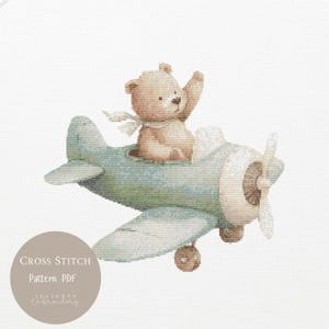 Könnte beinhalten: Ein Kreuzstichmuster mit einem Teddybären in einem hellblauen Flugzeug, der mit einem weißen Schal winkt. Das Flugzeug hat einen beigen Propeller und Räder. Der Text "CROSS STITCH Pattern PDF" befindet sich in einem Kreis. Ein charmantes Design für ein Kinderzimmer.