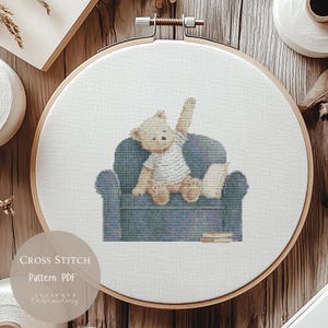 May include: A cross stitch pattern featuring a light brown teddy bear wearing a white striped shirt, sitting on a blue armchair with one arm raised. The bear is positioned next to a book, with two additional books resting on the floor by the chair.