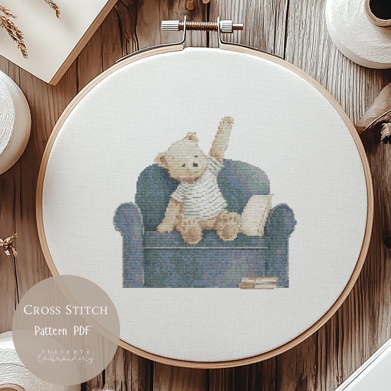 May include: A cross stitch pattern featuring a light brown teddy bear wearing a white striped shirt, sitting on a blue armchair with one arm raised. The bear is positioned next to a book, with two additional books resting on the floor by the chair.