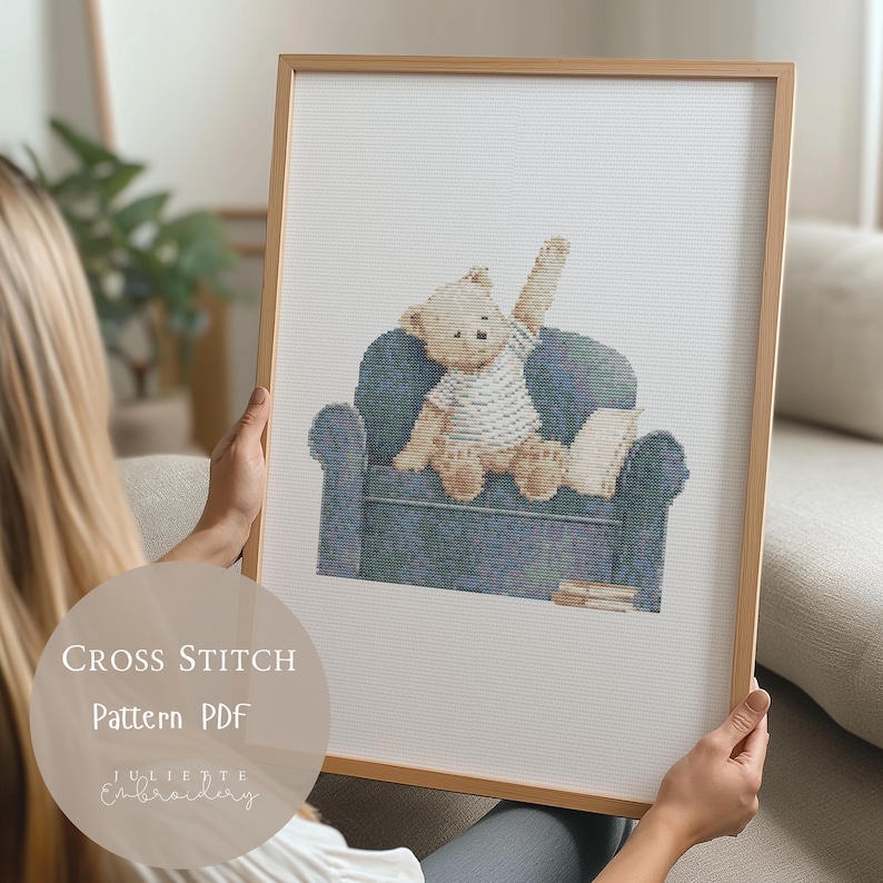 May include: A framed cross stitch pattern featuring a light brown teddy bear wearing a white striped shirt, sitting on a dark blue armchair with one arm raised. The bear is positioned on a white fabric background with a small book nearby.