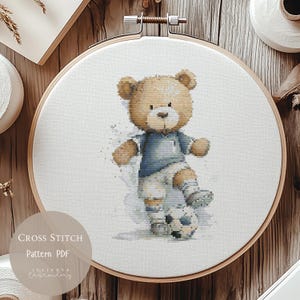 Puede incluir: Un patrón de punto de cruz con un oso de peluche con uniforme de fútbol pateando un balón. El oso es de color marrón claro, con una camiseta azul y pantalones cortos blancos. El diseño está en un bastidor de bordado de madera. Texto: "Cross Stitch Pattern PDF".