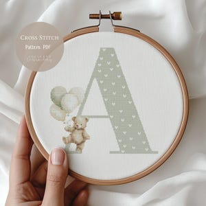 Può includere: Un motivo a punto croce in un telaio di legno presenta la lettera "A" in verde chiaro con un motivo a cuore. Un orsacchiotto tiene dei palloncini. Il testo "CROSS STITCH Pattern PDF" è in alto a sinistra.