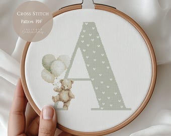 Può includere: Un motivo a punto croce in un telaio di legno presenta la lettera "A" in verde chiaro con un motivo a cuore. Un orsacchiotto tiene dei palloncini. Il testo "CROSS STITCH Pattern PDF" è in alto a sinistra.