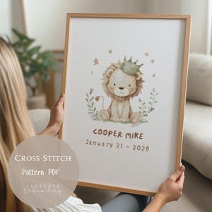 May include: A framed cross-stitch pattern featuring a cute lion wearing a crown, surrounded by leaves and stars. The text "COOPER MIKE" and "January 21 - 2026" are below the lion. The frame is light brown, and the background is white.