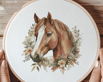 May include: A circular wooden embroidery hoop containing white fabric with a cross stitch design of a brown horse head surrounded by a wreath of muted green leaves and light-colored flowers. The text Cross Stitch Pattern PDF is visible.