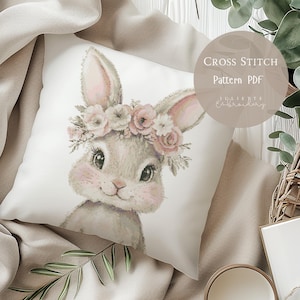 Puede incluir: Un cojín cuadrado blanquecino con un diseño de punto de cruz de un conejito con una corona floral. El conejito tiene pelaje gris, mejillas rosadas y ojos grandes y oscuros. El texto "Cross Stitch Pattern PDF" está en un círculo a la derecha.