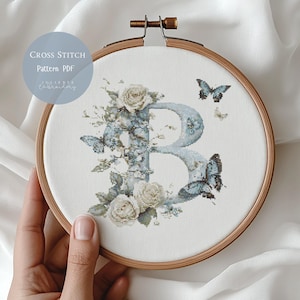 Peut inclure: Motif de point de croix avec la lettre "B" bleu clair, orné de roses blanches et de papillons bleus, dans un cercle à broder en bois. Le texte "Cross Stitch Pattern PDF" est également visible.