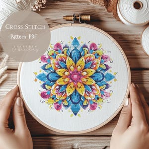 Könnte beinhalten: Ein farbenfrohes Kreuzstichmuster eines Mandalas in Blau-, Gelb-, Rosa- und Grüntönen. Das Muster ist in einem hölzernen Stickrahmen dargestellt. Der Text "Cross Stitch Pattern PDF" ist oben im Bild sichtbar.