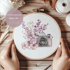 May include: Cross-stitch embroidery hoop with a vintage camera design and pink and purple floral accents. The text "Cross Stitch Pattern PDF" is visible. The embroidery is held in a wooden hoop.