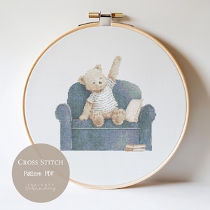May include: A cross stitch pattern in a wooden hoop featuring a light brown teddy bear wearing a white shirt, sitting on a blue armchair with one arm raised. Beside the bear is a book, and the text reads CROSS STITCH Pattern PDF JULIETTE EMBROIDERY.