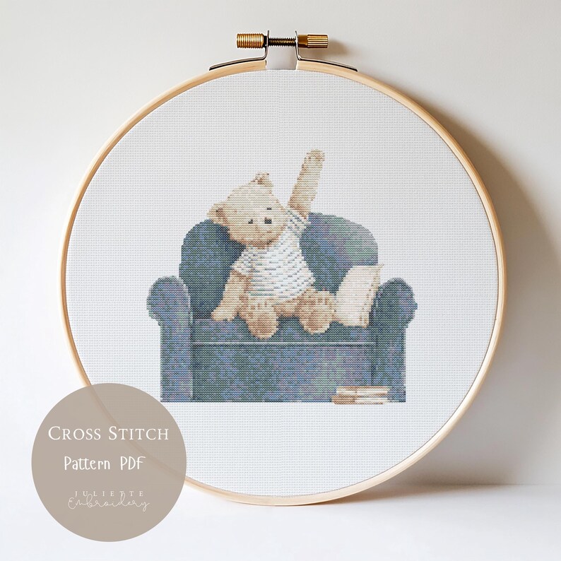 May include: A cross stitch pattern in a wooden hoop featuring a light brown teddy bear wearing a white shirt, sitting on a blue armchair with one arm raised. Beside the bear is a book, and the text reads CROSS STITCH Pattern PDF JULIETTE EMBROIDERY.