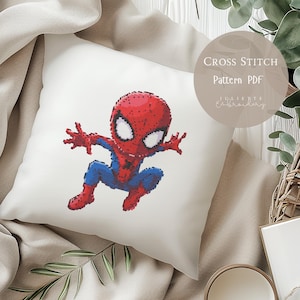 May include: A white square pillow features a cross-stitch design of Spider-Man in red and blue. The image includes the text "Cross Stitch Pattern PDF" and the name "Juliette Embroidery". The pillow is on a beige surface.