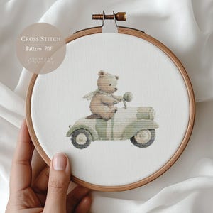 Könnte beinhalten: Kreuzstichmuster mit einem Teddybären, der einen Oldtimer-Roller fährt. Der Bär ist hellbraun mit einem grünen Schal, und der Roller ist hellgrün mit Streifen. Der Text "Cross Stitch Pattern PDF" befindet sich oben links.