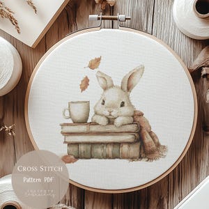 Könnte beinhalten: Ein Kreuzstichmuster mit einem weißen Kaninchen, das auf einem Stapel Bücher mit einer Tasse Kaffee sitzt. Das Kaninchen trägt einen braunen Schal und im Hintergrund fallen Blätter herunter. Der Text "CROSS STITCH Pattern PDF" befindet sich am unteren Rand des Bildes.