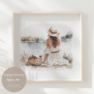 Puede incluir: Un patrón de punto de cruz que representa a una mujer con un vestido blanco y un sombrero de paja sentada junto a un lago. Ella sostiene una copa de vino y hay una cesta de fruta cerca. El patrón está en un marco blanco.