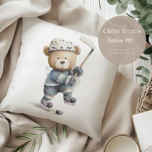 Op de afbeelding: Een wit kussen met een kruissteekontwerp van een teddybeer die hockey speelt. De beer draagt een blauw hockey-uniform en helm en houdt een hockeystick vast. De tekst "Cross Stitch Pattern PDF" is zichtbaar.