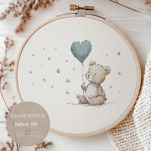 Puede incluir: Patrón de punto de cruz con un oso de peluche sosteniendo un globo en forma de corazón verde azulado. El diseño está en un bastidor de bordado de madera. El texto "Cross Stitch Pattern PDF" es visible.
