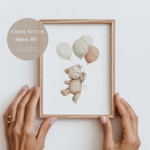 Op de afbeelding: Een ingelijst kruissteekpatroon met een teddybeer die ballonnen vasthoudt. De beer is lichtbruin en de ballonnen zijn pasteltinten roze, perzik en wit. De tekst "CROSS STITCH Pattern PDF" staat in een cirkel linksboven.