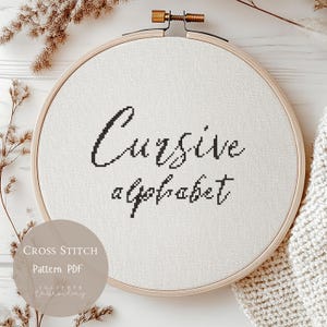 May include: A cross-stitch embroidery hoop displaying the words "Cursive alphabet" stitched in black thread on white fabric. The wooden hoop is round, and the text is in a handwritten style. A circular graphic with the words "Cross Stitch Pattern PDF" is in the lower left corner.