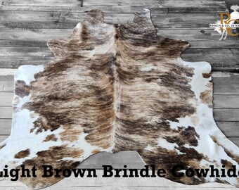 May include: Light brown brindle cowhide rug with white patches. The rug is laid out on a wooden floor.