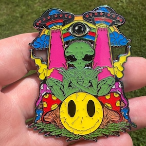 May include: A colorful enamel pin featuring a green alien with crossed arms, a yellow smiley face, and two UFOs. The pin has pink, blue, yellow, and red accents, with a psychedelic theme.