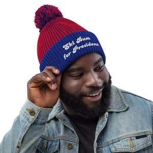 May include: A red and blue knit beanie with a matching pom-pom. The hat features the embroidered text "Ski Bum for President" in white. The beanie is worn by a person wearing a denim jacket.