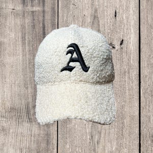 May include: Cream-colored baseball cap with a black embroidered letter "A". The cap is made of a soft, textured material and features a curved brim. The hat is a stylish accessory for casual wear.