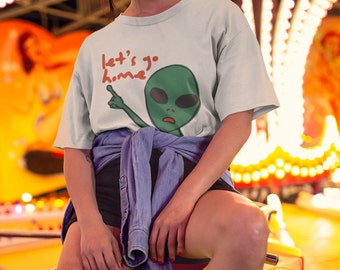 May include: A light gray t-shirt featuring a green alien graphic with the text "let's go home" in red. The alien is pointing with one hand. The shirt is part of a clothing collection.