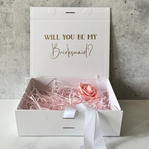 May include: White gift box with a pink rose inside. The lid of the box has the text "Will you be my Bridesmaid?" in gold lettering.